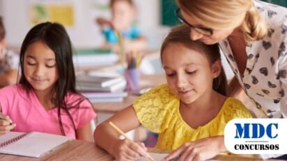 Uma professora com óculos e blusa branca estampada com corações auxilia uma aluna de blusa amarela em uma atividade escrita em sala de aula. A professora se inclina levemente sobre a mesa, apontando para o caderno da aluna, que escreve com um lápis laranja. Ao lado, outra estudante de blusa rosa também escreve em seu caderno. O ambiente é uma sala de aula iluminada, com materiais escolares como cadernos, lápis e livros sobre as mesas. Ao fundo, há um quadro verde e outras crianças desfocadas / Boa Vista - RR abre inscrições para Processo Seletivo com 325 Vagas e salários de até R$ 2,9 mil