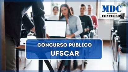 Um palestrante, vestindo um blazer escuro, está apoiado em uma mesa enquanto apresenta algo para um grupo de pessoas sentadas em cadeiras alinhadas. Na mesa, há um laptop aberto, um celular e outros itens. Os ouvintes, compostos por homens e mulheres, parecem atentos. O ambiente é uma sala iluminada por luz natural, com janelas ao fundo e algumas plantas decorativas / Concurso Público UFSCar 2025: 72 vagas para docentes com salários de até R$13,2 mil