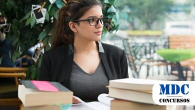 Mulher jovem de óculos e cabelos castanhos presos em um rabo de cavalo está sentada em uma mesa de café, cercada por livros. Ela veste um blazer preto sobre uma blusa cinza e olha pensativa para o lado, enquanto segura um livro aberto. O ambiente tem uma decoração aconchegante, com uma planta ao fundo adornada com fotos penduradas em prendedores de roupa. A janela ao lado reflete a movimentação da rua e permite a entrada de luz natural / Concurso em Capitão Leônidas Marques - PR oferece 29 vagas e salários de até R$20,1 mil