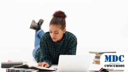 Mulher jovem de pele morena e cabelos cacheados presos em um coque está deitada no chão, vestindo uma camisa xadrez verde e preta, jeans e botas pretas. Ela está concentrada, lendo um livro aberto enquanto usa um laptop branco à sua frente. Ao redor dela, há vários livros empilhados e espalhados, além de um caderno com anotações. O fundo é branco, criando um ambiente minimalista e limpo / Concurso em Santa Maria Madalena - RJ oferece 147 vagas com salários de até R$ 5,6 mil