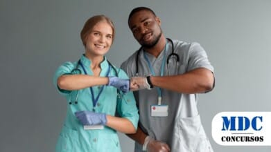 Processos Seletivos 6 Dois profissionais de saúde, um homem e uma mulher, usando jalecos, estetoscópios no pescoço e luvas, cumprimentam-se com toque de cotovelos enquanto sorriem, em um ambiente com fundo cinza / Consaúde VISA-CIS Vale do Aço – MG abre 8 vagas e salários de até R$4.750,00 em PSS