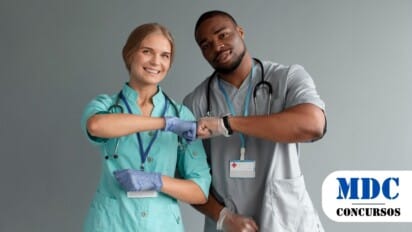 Dois profissionais de saúde, um homem e uma mulher, usando jalecos, estetoscópios no pescoço e luvas, cumprimentam-se com toque de cotovelos enquanto sorriem, em um ambiente com fundo cinza / Consaúde VISA-CIS Vale do Aço – MG abre 8 vagas e salários de até R$4.750,00 em PSS