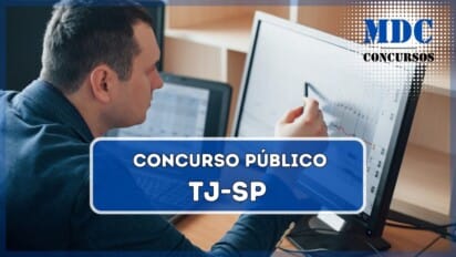 Um homem de pele clara e cabelos curtos escuros, vestindo um blazer azul, está sentado em uma mesa de escritório. Ele analisa gráficos exibidos em um monitor grande, segurando uma caneta para apontar detalhes na tela. Ao fundo, há um notebook aberto com outra tela exibindo gráficos e prateleiras vazias. / TJSP oferece 50 vagas para Analista de Sistemas com remuneração de até R$ 8.108,80
