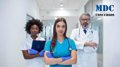 Três profissionais de saúde em um corredor de hospital. No centro, uma mulher jovem de cabelos longos e lisos, vestindo uniforme azul e estetoscópio rosa no pescoço, com os braços cruzados e expressão confiante. À esquerda, uma mulher com cabelo crespo, vestindo uniforme branco e segurando uma prancheta azul. À direita, um homem de cabelo grisalho e barba, vestindo jaleco branco, óculos e estetoscópio no pescoço, também com os braços cruzados. O ambiente é moderno e bem iluminado / HRAD de Patos de Minas abre Processo Seletivo em com Salários de até R$4.750,00