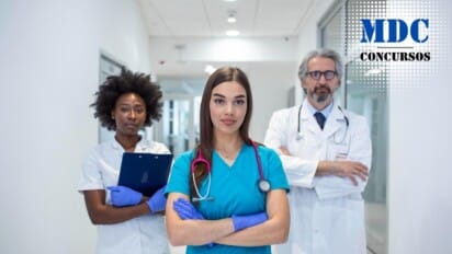 Três profissionais de saúde em um corredor de hospital. No centro, uma mulher jovem de cabelos longos e lisos, vestindo uniforme azul e estetoscópio rosa no pescoço, com os braços cruzados e expressão confiante. À esquerda, uma mulher com cabelo crespo, vestindo uniforme branco e segurando uma prancheta azul. À direita, um homem de cabelo grisalho e barba, vestindo jaleco branco, óculos e estetoscópio no pescoço, também com os braços cruzados. O ambiente é moderno e bem iluminado / HRAD de Patos de Minas abre Processo Seletivo em com Salários de até R$4.750,00