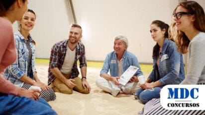 Grupo de seis pessoas sentadas em um círculo sobre a areia em um ambiente interno, parecendo participar de uma atividade descontraída. Um homem mais velho, de cabelo grisalho, segura uma prancheta e sorri enquanto conversa. Os outros participantes, de idades variadas, usam roupas casuais e demonstram envolvimento na conversa, com expressões alegres e atentas / Itamarati AM lança Processo Seletivo com 4 vagas e salários de até R$5 mil