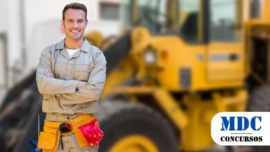 Processos Seletivos 8 Homem com uniforme bege de trabalho e cinto de ferramentas amarelo, sorrindo e de braços cruzados, em frente a uma máquina pesada amarela desfocada ao fundo. Ele carrega luvas vermelhas presas ao cinto e aparenta estar em um ambiente de construção ou manutenção / Maquiné – RS lança processo seletivo com 2 vagas e cadastro reserva para operador de máquinas