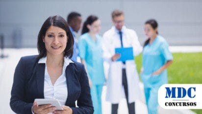 Mulher de terno preto e blusa branca segura um tablet e sorri para a câmera. Ao fundo, um grupo de profissionais de saúde, incluindo médicos e enfermeiros, conversam e analisam documentos. A cena ocorre em um ambiente externo moderno, com gramado e um prédio ao fundo / Oportunidade em Astolfo Dutra MG: Processo Seletivo com 7 Vagas e Salários de até R$3 mil