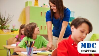 Processos Seletivos 7 Uma professora com blusa azul e calça bege está interagindo com um aluno de camiseta verde dentro de uma sala de aula. Ela se inclina levemente sobre a mesa, sorrindo e auxiliando o estudante, que a olha com atenção. Em primeiro plano, outro aluno de camisa vermelha está escrevendo em um caderno. Ao fundo, uma aluna de blusa rosa também está concentrada em sua atividade. A sala de aula tem móveis de madeira clara, armários verdes e elementos decorativos coloridos / PSS em Paulistana - PI: Mais de 50 vagas na área da Educação e Assistência Social