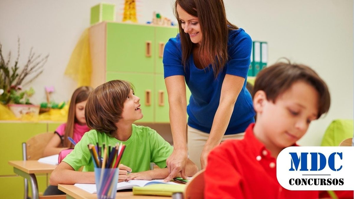Uma professora com blusa azul e calça bege está interagindo com um aluno de camiseta verde dentro de uma sala de aula. Ela se inclina levemente sobre a mesa, sorrindo e auxiliando o estudante, que a olha com atenção. Em primeiro plano, outro aluno de camisa vermelha está escrevendo em um caderno. Ao fundo, uma aluna de blusa rosa também está concentrada em sua atividade. A sala de aula tem móveis de madeira clara, armários verdes e elementos decorativos coloridos / PSS em Paulistana - PI: Mais de 50 vagas na área da Educação e Assistência Social