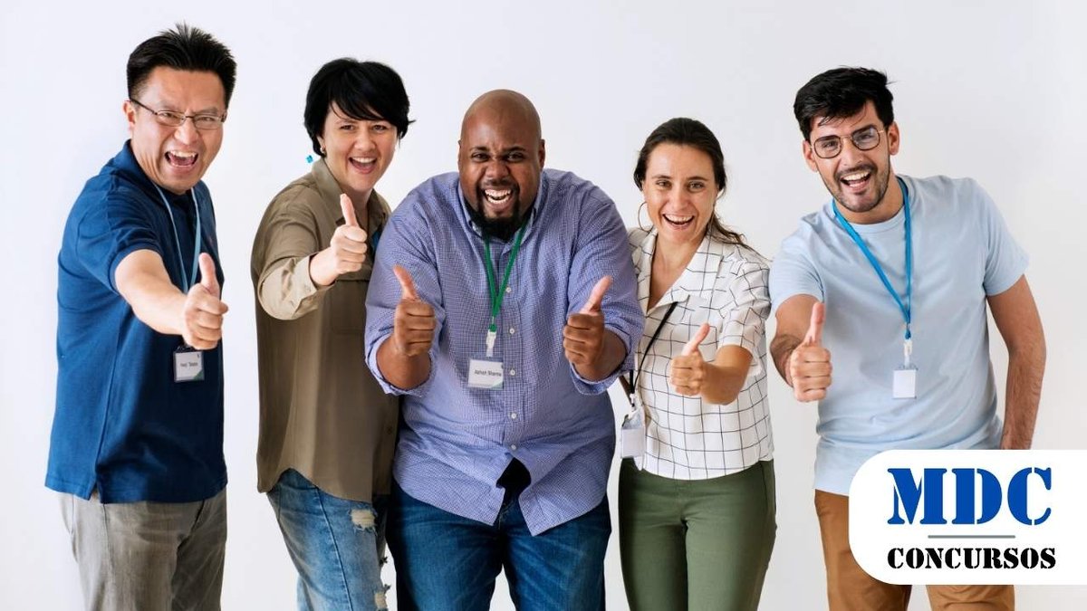 Grupo de cinco pessoas diversas, sorridentes e demonstrando entusiasmo com polegares para cima. Todos usam crachás e estão em um ambiente claro e neutro. Eles parecem estar em um evento corporativo ou equipe de trabalho, transmitindo energia positiva e colaboração / Prefeitura de Apodi - RN lança processos seletivos com 32 vagas em diferentes Secretarias