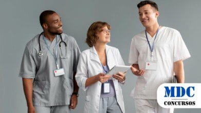 Processos Seletivos 9 Três profissionais de saúde conversam sorrindo. Uma médica, vestindo jaleco branco e segurando um tablet, está no centro. À esquerda, um enfermeiro de uniforme cinza com estetoscópio no pescoço. À direita, outro profissional de saúde veste uniforme branco e segura uma prancheta. Todos usam crachás e parecem engajados na conversa / Prefeitura de Estiva abre inscrições para contratação na área da Saúde