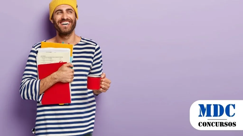 Homem sorridente usando gorro amarelo e camiseta listrada azul e branca, segurando uma caneca vermelha e uma pilha de pastas e papéis coloridos, em frente a uma parede lilás / Prefeitura de Jaguapitã PR lança Processo Seletivo com salários de R$6 mil