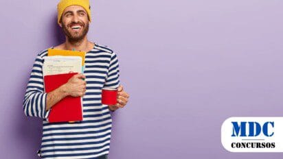Homem sorridente usando gorro amarelo e camiseta listrada azul e branca, segurando uma caneca vermelha e uma pilha de pastas e papéis coloridos, em frente a uma parede lilás / Prefeitura de Jaguapitã PR lança Processo Seletivo com salários de R$6 mil