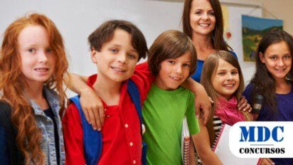 Grupo de crianças sorridentes com mochilas e cadernos ao lado de uma professora em uma sala de aula. Elas parecem felizes e próximas, transmitindo um ambiente amigável e educativo. A professora, ao fundo, usa uma blusa azul e sorri, enquanto as crianças vestem roupas coloridas e demonstram interação entre si / Recreio MG 2025: Processo Seletivo oferece vaga + Cadastro de Reserva