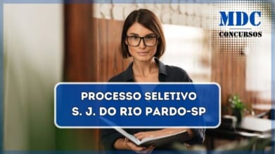 Mulher jovem com cabelos curtos castanhos e óculos de armação preta, vestindo uma camisa escura, segurando um caderno aberto e olhando diretamente para a câmera com uma expressão confiante. O ambiente ao fundo tem uma parede com revestimento de madeira e tijolos brancos, além de móveis e plantas, sugerindo um espaço moderno e aconchegante / Prefeitura de São José do Rio Pardo - SP abre Processo Seletivo com salário de R$ 6.905,41
