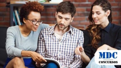 Três pessoas jovens em um sofá, imersas na leitura de um livro, simbolizando a troca de conhecimento e companheirismo / Prefeitura de Tremembé - SP lança concurso com 126 vagas + CR e salários de até R$4,9 mil