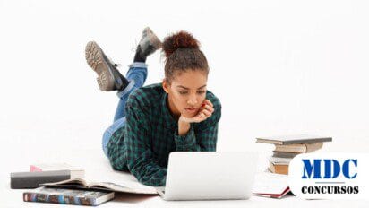 Jovem deitada no chão, usando camisa xadrez verde e jeans, concentrada em um laptop. Livros abertos e empilhados estão ao redor dela, em um ambiente de fundo branco / Processo Seletivo Afrânio - PE 417 vagas e salários de até R$ 10,5 mil