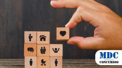 Processos Seletivos 9 Uma mão segurando um bloco de madeira com um ícone de coração, posicionando-o no topo de uma pilha de outros blocos de madeira. Os blocos abaixo contêm ícones representando diferentes aspectos da vida, como moradia, família, comunicação, saúde, gravidez, e crianças. O fundo é escuro e desfocado, enquanto a cena está sobre uma superfície de madeira / Processo Seletivo Hidrolândia - GO 35 vagas e salários de até R$ 3,1 mil