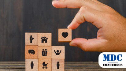 Uma mão segurando um bloco de madeira com um ícone de coração, posicionando-o no topo de uma pilha de outros blocos de madeira. Os blocos abaixo contêm ícones representando diferentes aspectos da vida, como moradia, família, comunicação, saúde, gravidez, e crianças. O fundo é escuro e desfocado, enquanto a cena está sobre uma superfície de madeira / Processo Seletivo Hidrolândia - GO 35 vagas e salários de até R$ 3,1 mil