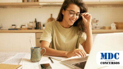 Mulher sorridente com óculos e camiseta bege trabalha em um laptop na cozinha. Na mesa, há papéis, um caderno, caneta, celular e uma caneca azul. O ambiente tem tons neutros e iluminação natural / Processo Seletivo União de Minas MG: 4 vagas para profissionais de nível superior