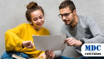 Dois jovens sentados no chão contra uma parede cinza, analisando juntos uma folha de papel. A pessoa à esquerda, com cabelo preso em um coque e vestindo um suéter amarelo, sorri enquanto aponta para o documento. A pessoa à direita, usando óculos, camiseta cinza e relógio preto, também observa a folha com atenção. Há um livro aberto no colo da pessoa à esquerda. Ambos parecem estar estudando ou trabalhando em um projeto colaborativo / Processo Seletivo de Ouro Verde – SP oferece oportunidades p/ níveis médio e superior