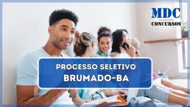 Uma imagem de um grupo de jovens sentados no chão de um corredor iluminado, encostados em uma parede branca. O foco está em um jovem sorridente de camiseta azul clara, segurando uma caneta e um caderno aberto. Atrás dele, quatro jovens estão envolvidos em diferentes atividades, como conversar, usar um laptop e escrever em cadernos. Eles parecem estar estudando ou discutindo algo em um ambiente acadêmico ou universitário. O local tem uma atmosfera descontraída e colaborativa, com fones de ouvido, laptops e materiais de estudo espalhados / Processo Seletivo em Brumado  - BA abre 80 Vagas no Sistema Municipal de Ensino