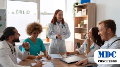 Grupo de profissionais da área da saúde em uma reunião em um ambiente de escritório iluminado. Cinco pessoas estão reunidas ao redor de uma mesa de madeira, analisando gráficos e documentos. Duas mulheres vestem jalecos brancos, uma delas em pé, falando e sorrindo para o grupo, enquanto as outras três pessoas, incluindo um homem e uma mulher com uniforme cirúrgico verde, estão sentadas, ouvindo e interagindo. Há um notebook sobre a mesa e uma estante com plantas e caixas organizadoras ao fundo. A atmosfera é descontraída e colaborativa / Processo Seletivo em Buritis - MG: 8 vagas e salários de até R$6.500,00