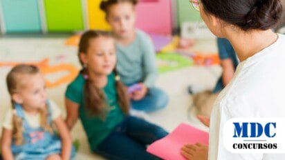 Uma professora, vestindo uma camiseta branca e óculos, está sentada no chão de uma sala colorida de educação infantil, segurando uma folha rosa enquanto conversa com um grupo de três meninas. As crianças, que parecem ter entre 4 e 7 anos, estão sentadas no tapete e prestam atenção à professora. Ao fundo, há caixas organizadoras coloridas e materiais escolares espalhados pelo chão, sugerindo um ambiente educativo e lúdico / Processo Seletivo em Mâncio Lima AC oferece 49 vagas e salários de até R$ 3,4 mil