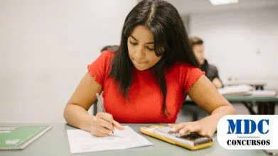 Mulher jovem de cabelo longo e escuro, vestindo camiseta vermelha, sentada em uma carteira escolar, concentrada enquanto escreve em uma folha de papel e usa uma calculadora / Processo Seletivo em Monte Negro - RO oferece 20 vagas e salários de até R$ 7 mil