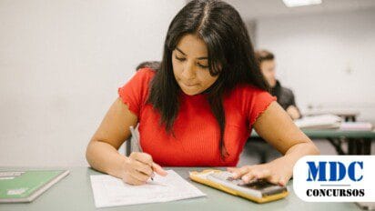 Mulher jovem de cabelo longo e escuro, vestindo camiseta vermelha, sentada em uma carteira escolar, concentrada enquanto escreve em uma folha de papel e usa uma calculadora / Processo Seletivo em Monte Negro - RO oferece 20 vagas e salários de até R$ 7 mil