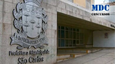 Fachada do prédio da Prefeitura Municipal de São Carlos, com destaque para o brasão do município em relevo na parede de pedra. O brasão contém o lema em latim "A BANDEIRANTIBUS VENIO". À direita, há uma entrada coberta com rampa de acesso e janelas de vidro / Processo Seletivo em São Carlos - SP oferece salário de R$ 3 mil p/ Agente de Combate às Endemias