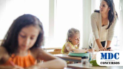 Uma professora jovem, de cabelos castanhos longos, veste um cardigã bege sobre uma blusa branca e está inclinada sobre uma mesa de sala de aula, observando e auxiliando duas alunas. Uma menina loira, usando um vestido amarelo, está concentrada escrevendo em um caderno, enquanto outra menina, de cabelos escuros e vestindo uma blusa laranja, aparece desfocada no primeiro plano também escrevendo. A sala de aula é iluminada por luz natural vinda de grandes janelas ao fundo, e há materiais escolares espalhados sobre as mesas / Processo Seletivo para Professores em Santo Antônio RN oferece 18 Vagas