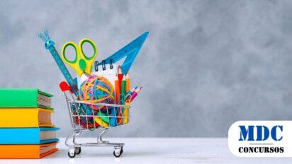 Carrinho de compras em miniatura cheio de materiais escolares coloridos, incluindo lápis, tesoura, régua e borrachas, ao lado de uma pilha de livros de capa colorida. O fundo é cinza texturizado e a mesa branca, criando um contraste vibrante com os itens escolares / SEMED Coari AM oferece 181 vagas para apoio à educação com salários de até R$ 3 mil