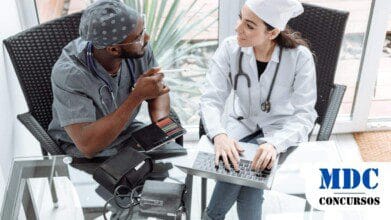 Processos Seletivos 2 Dois profissionais da saúde estão sentados à mesa em um ambiente iluminado e moderno. Um deles, um homem negro vestindo uniforme cinza, touca cirúrgica e estetoscópio no pescoço, segura um tablet com lápis de cor e gesticula com uma caneta. A outra pessoa, uma mulher de pele clara vestindo jaleco branco, touca cirúrgica e estetoscópio, digita em um laptop enquanto sorri para seu colega. Na mesa de vidro há um esfigmomanômetro (medidor de pressão arterial) e outros materiais médicos. O ambiente possui paredes de vidro, permitindo a entrada de luz natural e uma vista para um espaço externo com plantas / Santa Vitória - MG lança PSS na área da saúde com salários de até R$ 4,1 mil