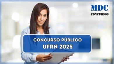 Uma mulher com cabelo castanho longo e pele clara está segurando um caderno de capa espiral aberta e olhando diretamente para a câmera com uma expressão confiante e séria. Ela veste uma camisa social azul clara e está em um ambiente bem iluminado, que parece ser um escritório moderno ou uma sala de reunião. O fundo está desfocado, com tons claros e elementos sutis de móveis e iluminação / UFRN lança editais de Concurso Público com salários de até R$ 4.967,04