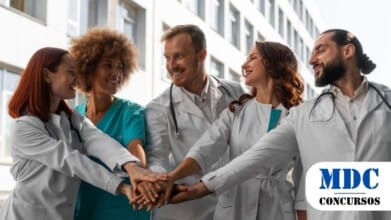 Processos Seletivos 2 Grupo de cinco profissionais de saúde, usando jalecos brancos e roupas médicas, reunidos em um gesto de união com as mãos empilhadas ao centro. Eles sorriem e se olham, demonstrando espírito de equipe e colaboração. Ao fundo, há um edifício moderno com janelas grandes, sugerindo o ambiente hospitalar ou clínico / UNIMONTES oferece 58 vagas em Programas de Residência com bolsas de até R$ 4.106,09