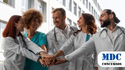 Grupo de cinco profissionais de saúde, usando jalecos brancos e roupas médicas, reunidos em um gesto de união com as mãos empilhadas ao centro. Eles sorriem e se olham, demonstrando espírito de equipe e colaboração. Ao fundo, há um edifício moderno com janelas grandes, sugerindo o ambiente hospitalar ou clínico / UNIMONTES oferece 58 vagas em Programas de Residência com bolsas de até R$ 4.106,09