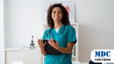 Jovem profissional da saúde com cabelos cacheados e uniforme verde segurando uma prancheta e sorrindo em um consultório médico. Ao fundo, há cartazes anatômicos, um microscópio e equipamentos médicos. O ambiente é moderno e bem iluminado, transmitindo uma sensação de profissionalismo e acolhimento / Alumínio (SP) Lança Processo Seletivo