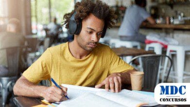 Homem jovem de camiseta amarela e fones de ouvido grandes estudando em um café. Ele escreve em um caderno enquanto consulta um livro aberto. Há anotações, um celular e um copo descartável sobre a mesa. O ambiente tem luz natural, móveis rústicos e pessoas ao fundo - Anajás (PA) lança Processo Seletivo com 91 Vagas