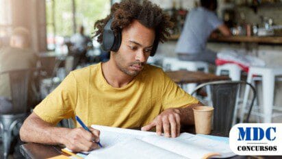 Homem jovem de camiseta amarela e fones de ouvido grandes estudando em um café. Ele escreve em um caderno enquanto consulta um livro aberto. Há anotações, um celular e um copo descartável sobre a mesa. O ambiente tem luz natural, móveis rústicos e pessoas ao fundo - Anajás (PA) lança Processo Seletivo com 91 Vagas