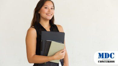 Processos Seletivos 6 Mulher jovem sorridente com cabelos longos e castanhos segurando cadernos e uma pasta preta contra um fundo branco, vestindo uma blusa preta sem mangas e calça branca - Atalaia do Norte (AM) Abre Inscrições para 300 Vagas em Estágio