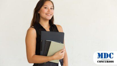 Mulher jovem sorridente com cabelos longos e castanhos segurando cadernos e uma pasta preta contra um fundo branco, vestindo uma blusa preta sem mangas e calça branca - Atalaia do Norte (AM) Abre Inscrições para 300 Vagas em Estágio