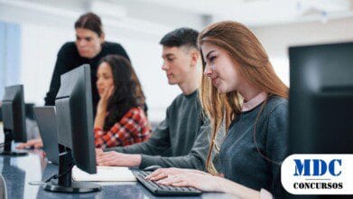Jovens em uma sala de informática estudando em computadores. Uma jovem de cabelos longos digita no teclado sorrindo. Ao fundo, um professor observa os alunos. No canto inferior direito, há um logotipo com o texto "MDC CONCURSOS" em azul e preto - Banrisul abre 100 Vagas com Salário Inicial de R$ 5 mil em Concurso na Área de TI