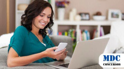 Mulher sorridente de blusa verde sentada em um sofá usando um notebook e um celular, aparentando estudar ou trabalhar. Ao fundo, uma estante com livros e itens decorativos. No canto inferior direito, o logotipo “MDC Concursos” indica relação com concursos públicos ou educação - Borebi (SP) Abre Concurso com 10 Vagas e Salários de até R$ 4,5 mil