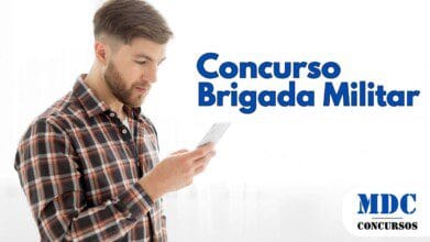 Homem jovem com barba e cabelo castanho curto, vestindo camisa xadrez, olhando para seu smartphone com expressão concentrada. O fundo é branco e iluminado, criando um contraste com sua roupa escura - Brigada Militar Abre Concurso com 1.200 Vagas e Salário de R$ 5.944,85