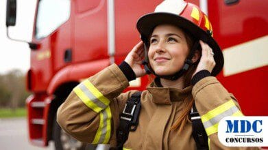 Processos Seletivos 2 Mulher bombeira com uniforme bege e faixas refletivas ajusta o capacete vermelho com detalhes amarelos. Ela sorri confiante, com um caminhão de bombeiros vermelho ao fundo / CBMMT lança Processo Seletivo para a área da Saúde com salários de até R$ 6,5 mil