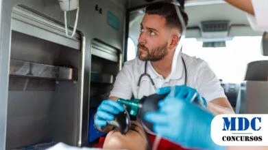 Profissional de saúde dentro de uma ambulância realizando atendimento de emergência com um ressuscitador manual (Ambu). Ele usa luvas azuis e uniforme branco, demonstrando concentração. O ambiente possui equipamentos médicos. No canto inferior direito, há um logotipo com os dizeres "MDC CONCURSOS" / CONSAMU PR abre Processo Seletivo com salários de até R$ 6,4 mil e 61 vagas