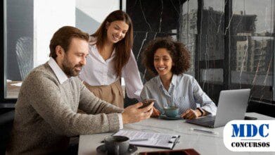 Três colegas de trabalho em um ambiente corporativo moderno. Um homem barbudo de suéter segura um smartphone, enquanto duas mulheres, uma de cabelos cacheados e outra de cabelos lisos, sorriem e observam. Há um laptop, café e documentos na mesa. O ambiente tem janelas amplas com vista para prédios urbanos e paredes de mármore preto / Concurso CORE-BA