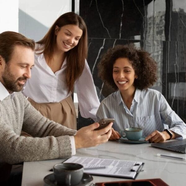 Três colegas de trabalho em um ambiente corporativo moderno. Um homem barbudo de suéter segura um smartphone, enquanto duas mulheres, uma de cabelos cacheados e outra de cabelos lisos, sorriem e observam. Há um laptop, café e documentos na mesa. O ambiente tem janelas amplas com vista para prédios urbanos e paredes de mármore preto / Concurso CORE-BA
